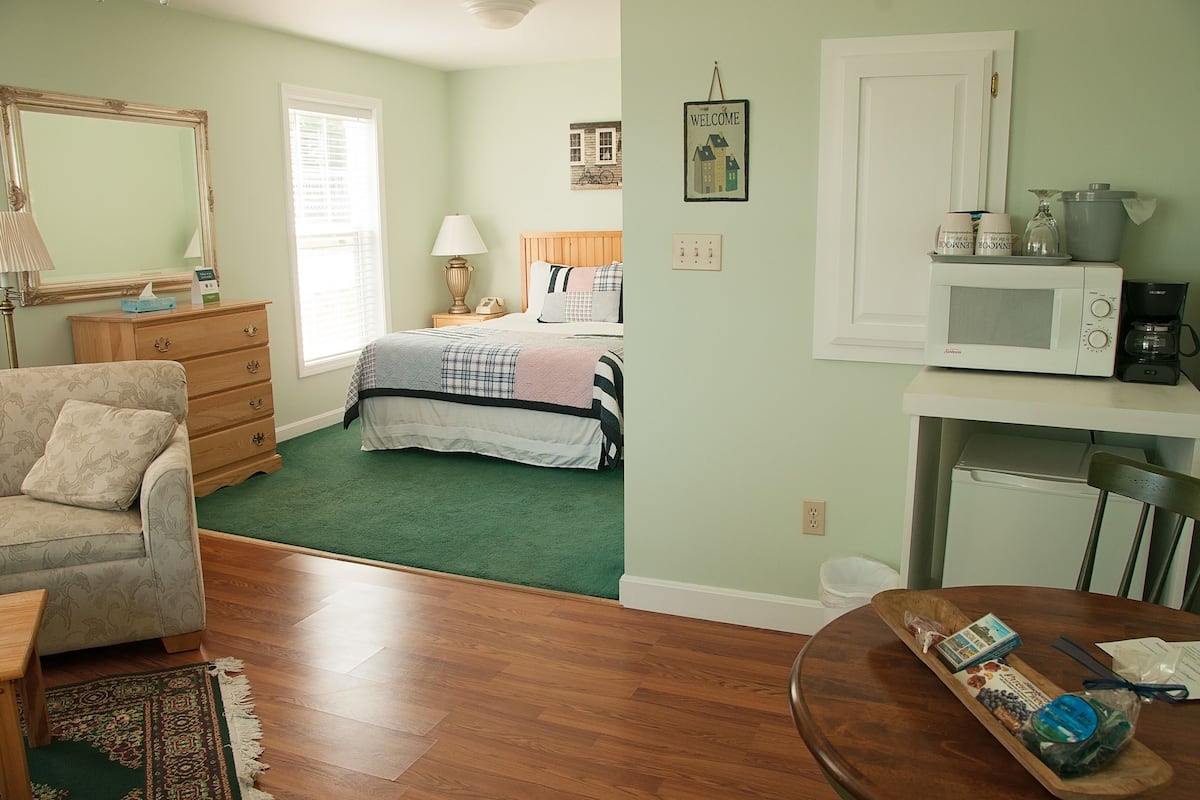 A bedroom with a bed, dresser, and a nightstand with a lamp. There is a dining area with a table and chairs.