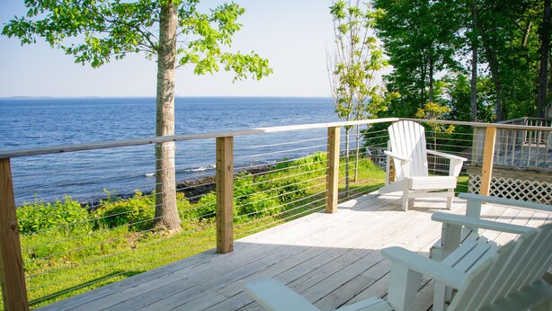 A wooden deck with white Adirondack chairs overlooking a body of water. - Glenmoor By the Sea (Lincolnville)