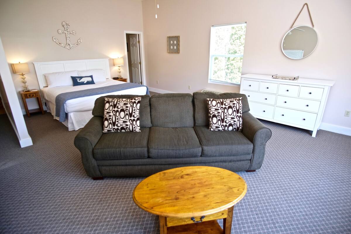 A hotel room with a bed, a gray sofa with patterned cushions, a wooden coffee table, a white dresser, and a wall-mounted mirror.