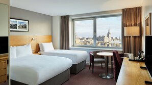 A hotel room with two beds, a desk, a chair, a small table, and a large window offering a city view.