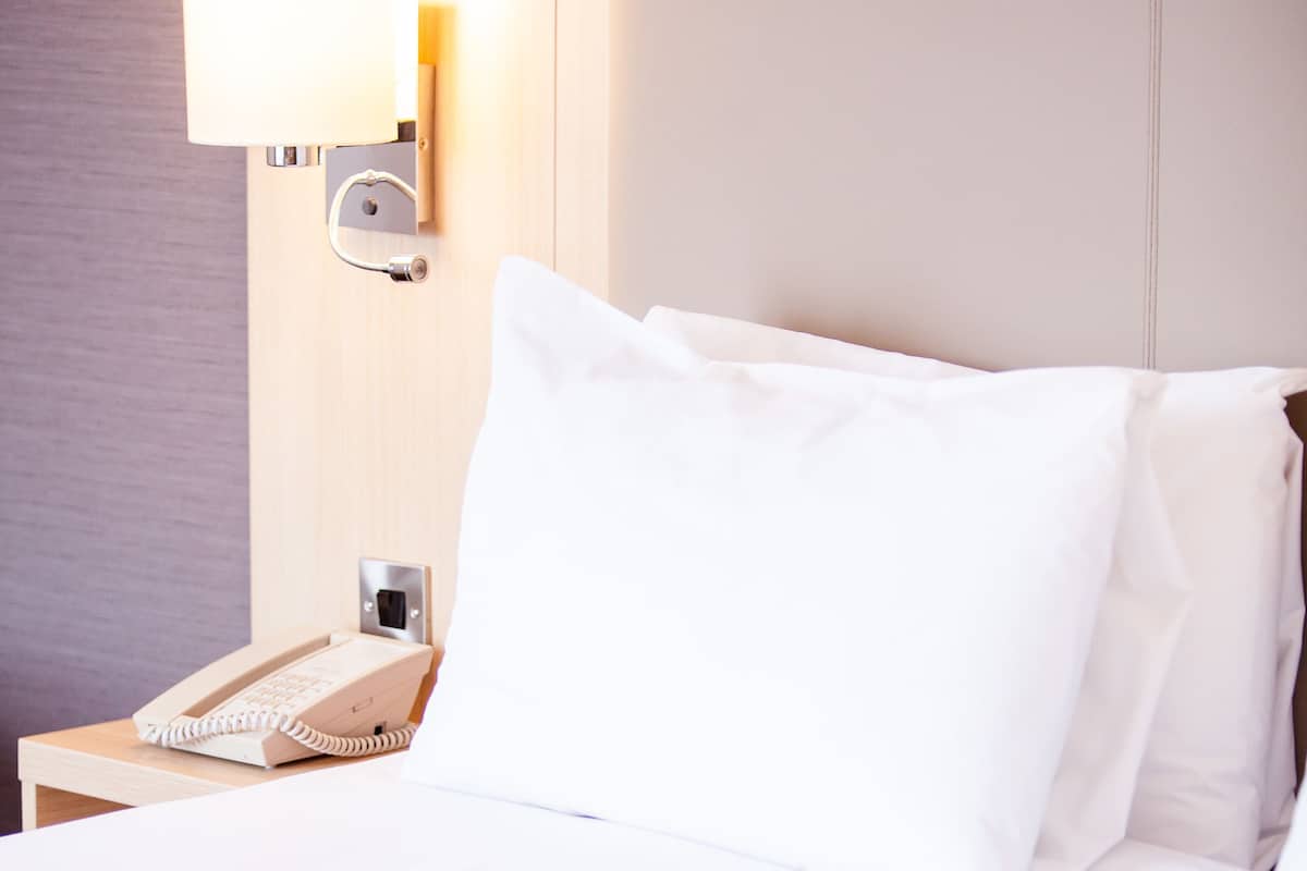 Uma cama bem arrumada com lençóis brancos, um telefone ao lado e uma luminária na parede.