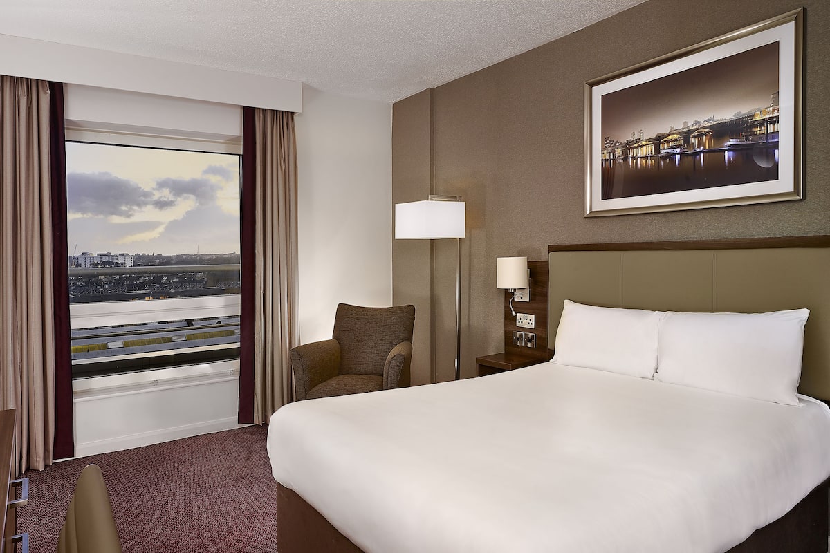 Quarto de hotel com uma cama grande, uma mesa de trabalho com uma TV, uma cadeira, uma mesa de cabeceira com abajur e uma janela com cortinas.
