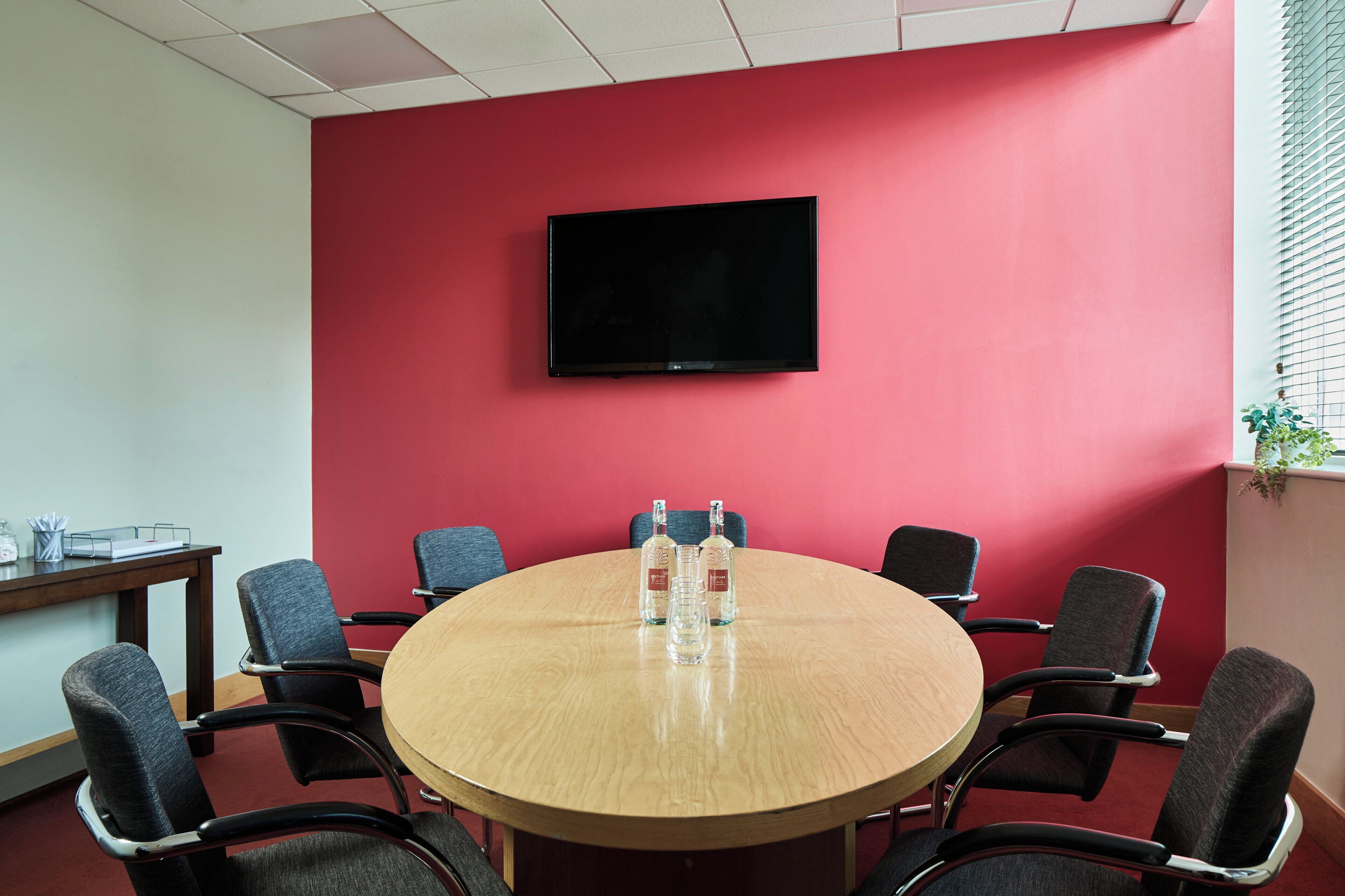 A conference room with a round table, six chairs, a flat-screen TV on the wall, and two bottles on the table.