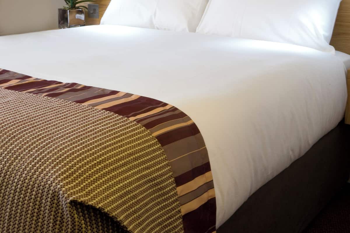 Uma cama bem arrumada com lençóis brancos, cabeceira de madeira e um manta decorativa.