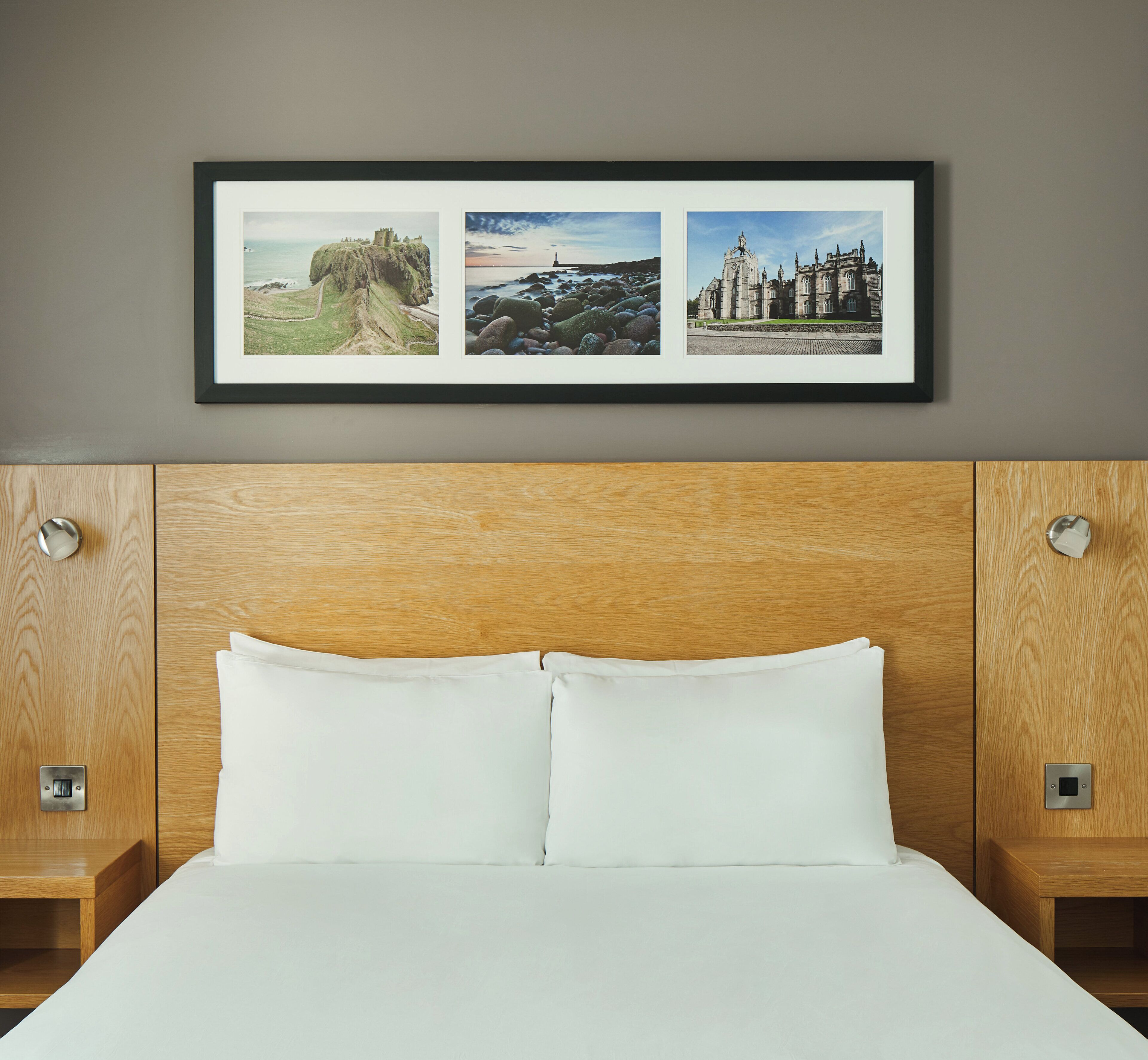 Habitación de hotel con cabecera de madera, dos almohadas blancas y un cuadro en la pared con un castillo, un faro y una catedral.