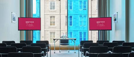 A conference room with a table and chairs, two screens displaying 'MEETINGS AND EVENTS' for the LEONARDO Hotels, and a view of a blue building through the windows.
