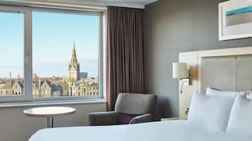 Ein Hotelzimmer mit einem großen Bett, einem Sessel, einem kleinen Tisch und Blick auf eine Stadtlandschaft mit einem Kirchturm.