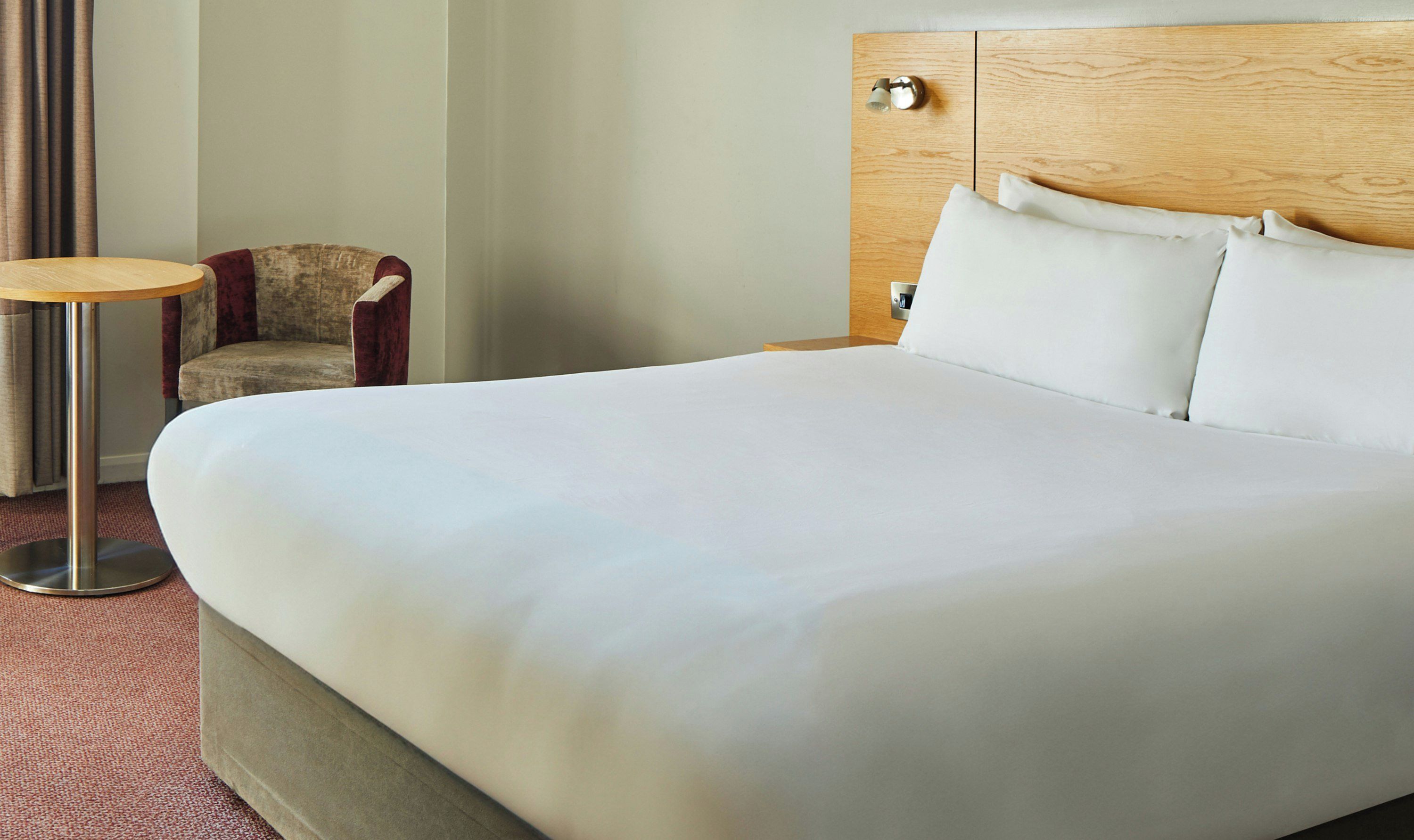 Une chambre d’hôtel avec un grand lit, une tête de lit en bois, une petite table ronde et un fauteuil rouge.