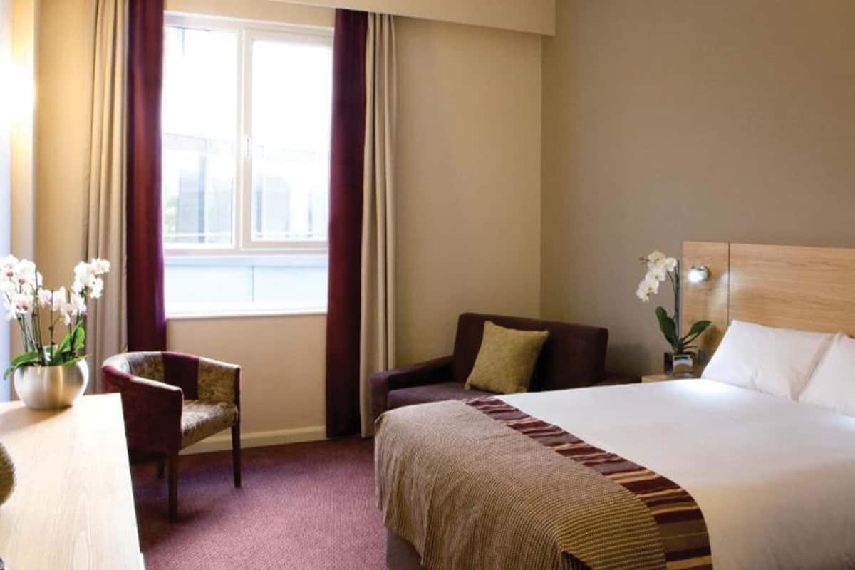 Quarto de hotel com cama, uma cadeira, escrivaninha e janela com cortinas.