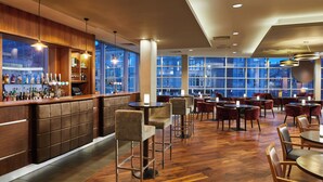 A modern bar area with a wooden bar counter, multiple seating options including high stools and round tables, and large windows offering a city view.