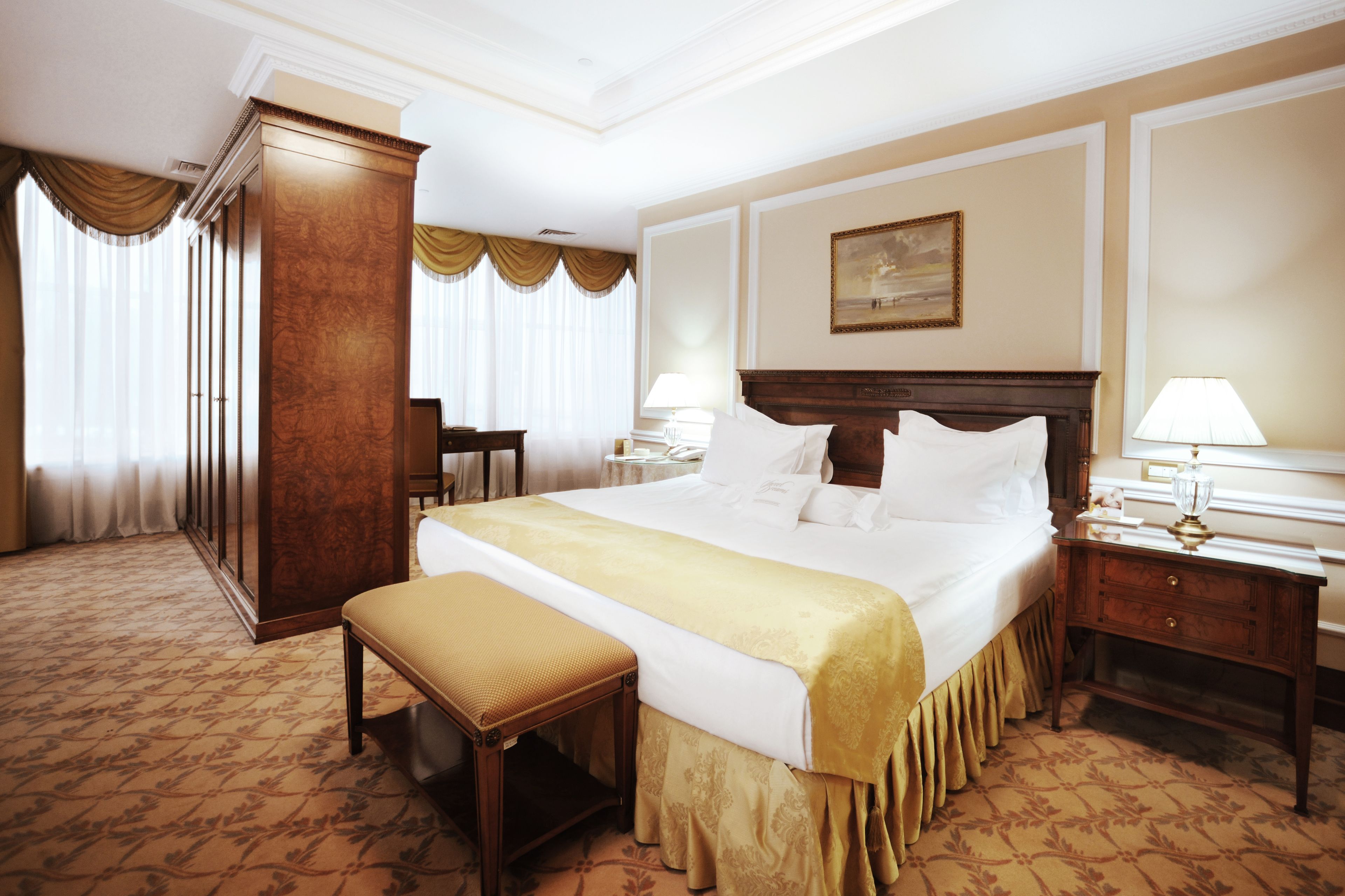 A hotel room with a large bed, a wooden wardrobe, a bench, a nightstand, and a lamp.