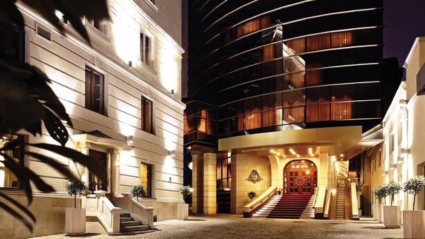 Una entrada de hotel con una imponente escalera, un edificio moderno de fachada curva y un patio adoquinado.