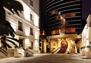 Una entrada de hotel con una imponente escalera, un edificio moderno de fachada curva y un patio adoquinado.