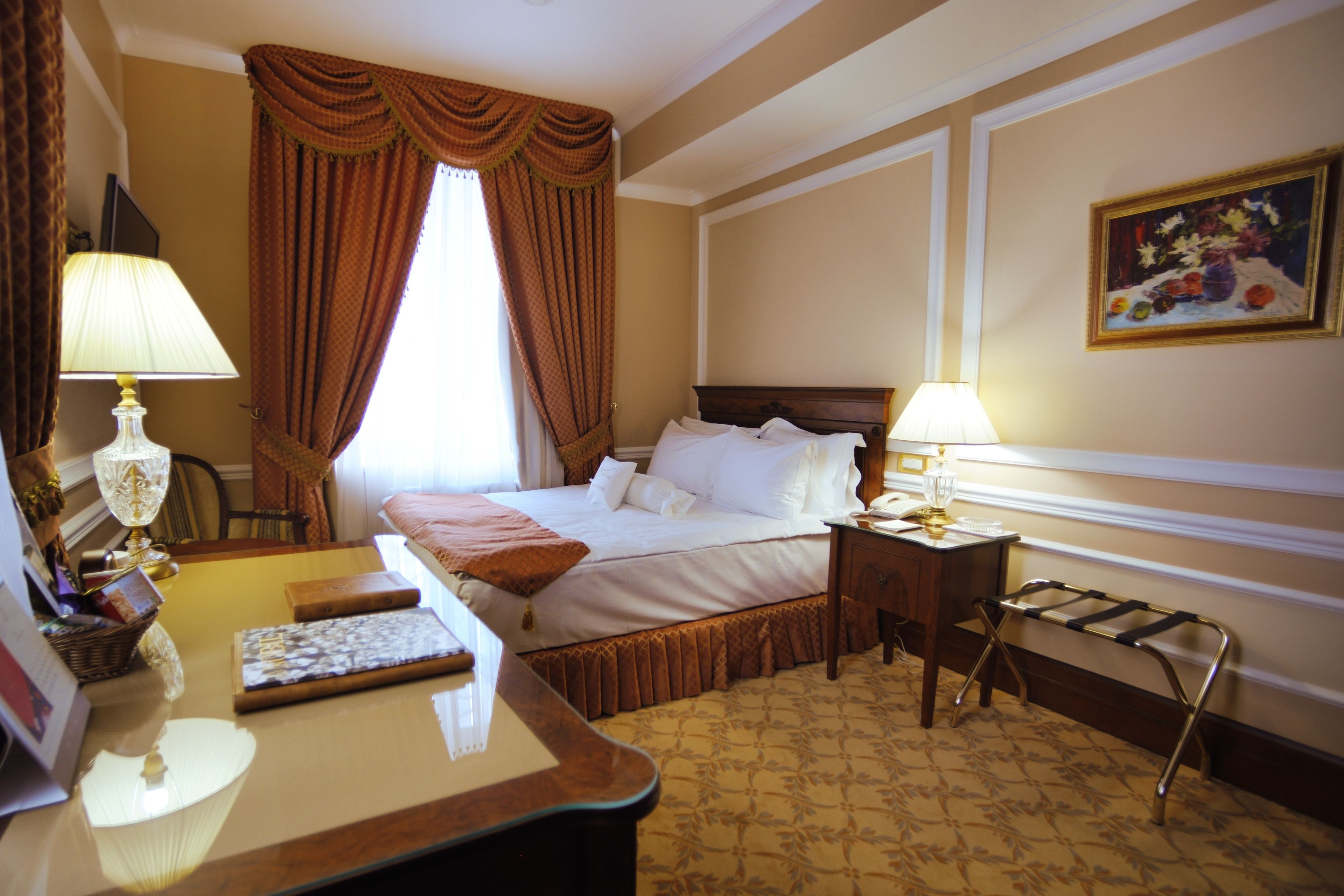 A hotel room with a bed, two bedside tables, a desk with a lamp, a chair, a small table with a lamp, and a painting on the wall.