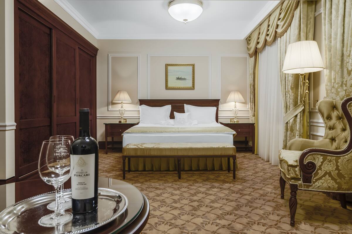 Habitación de hotel con una cama grande, dos mesitas de noche con lámparas, un banco de madera, una silla decorativa y una botella de vino sobre una bandeja.