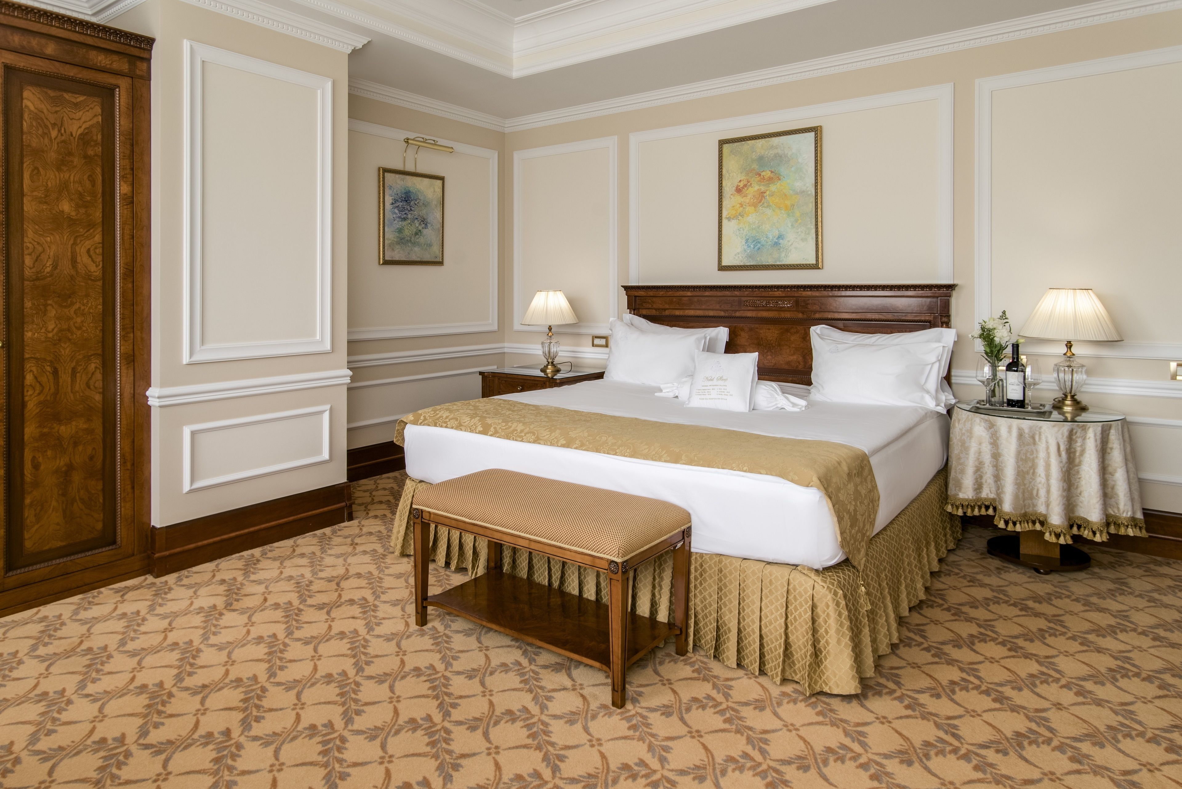 A hotel room with a large bed, a wooden bench, two bedside tables with lamps, and a large wooden wardrobe.