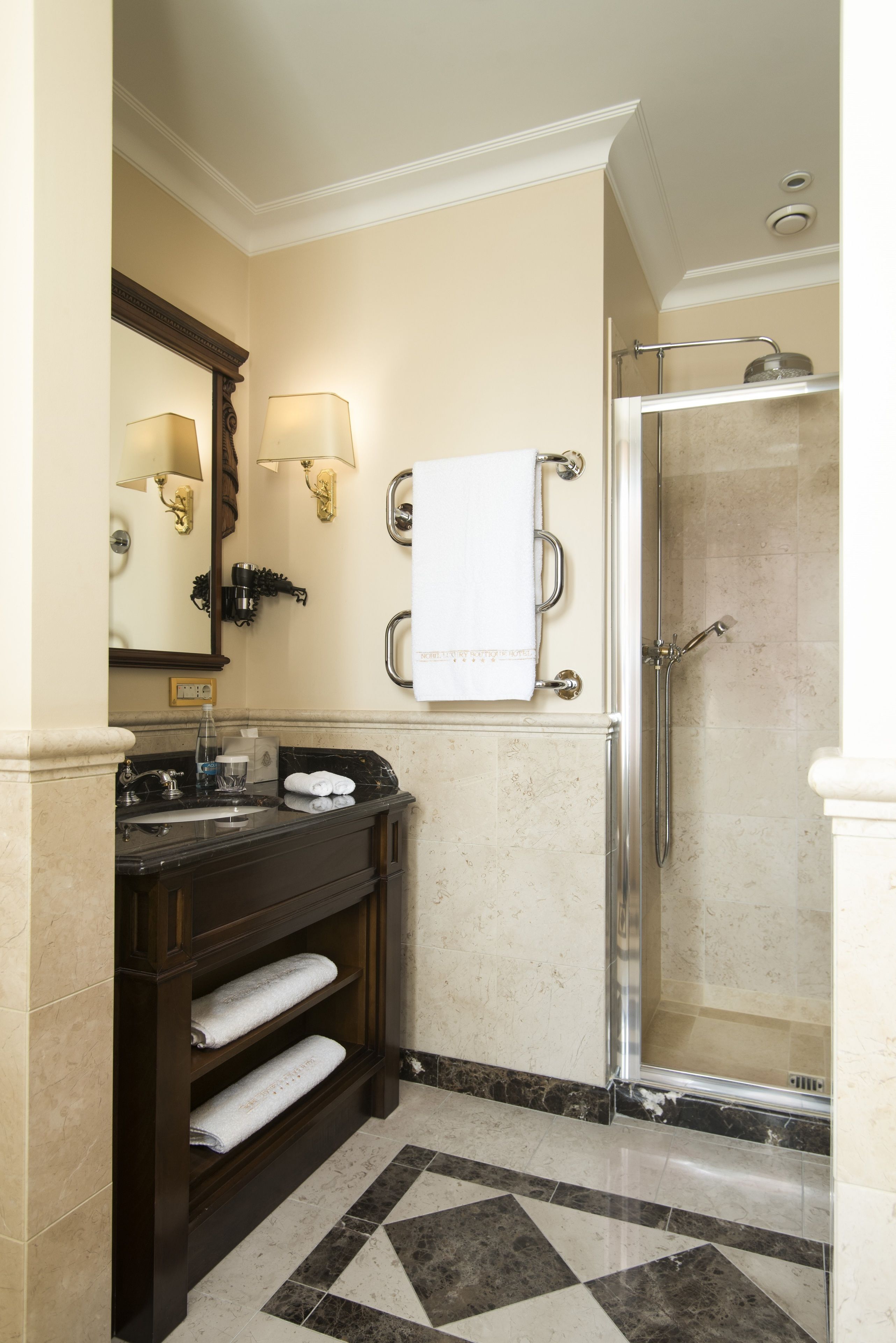 A bathroom with a glass-enclosed shower, a dark wooden vanity with a mirror and two wall-mounted lights, and a tiled floor with a geometric pattern.
