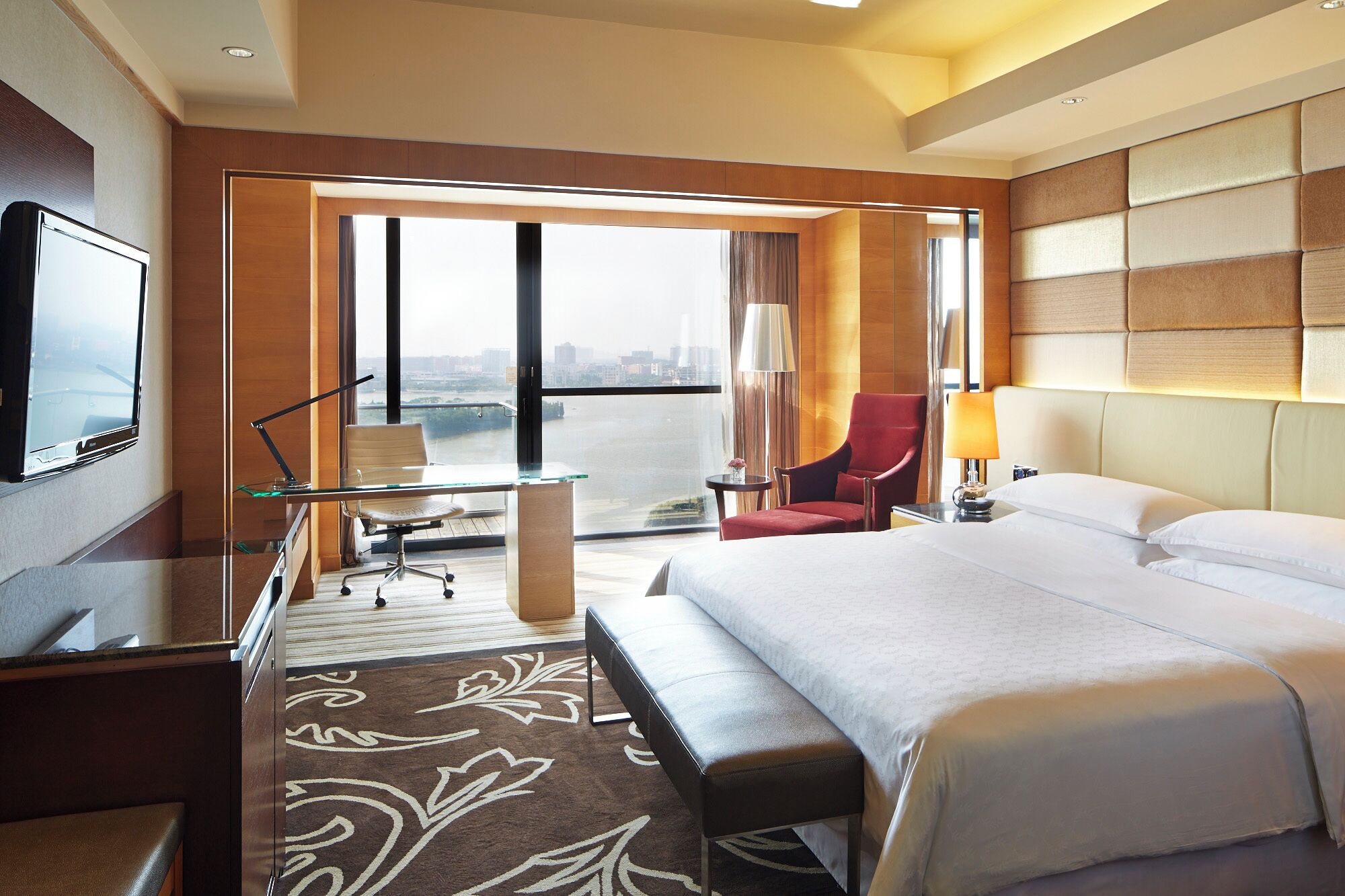A hotel room with a bed, a desk with a chair, a red armchair, a TV, and a view of a cityscape through large windows.
