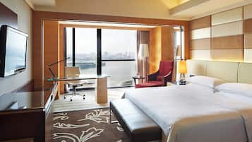Ein Hotelzimmer mit einem Bett, einem Schreibtisch mit Stuhl, einem roten Sessel, einem Fernseher und Blick auf eine Stadtlandschaft durch große Fenster.