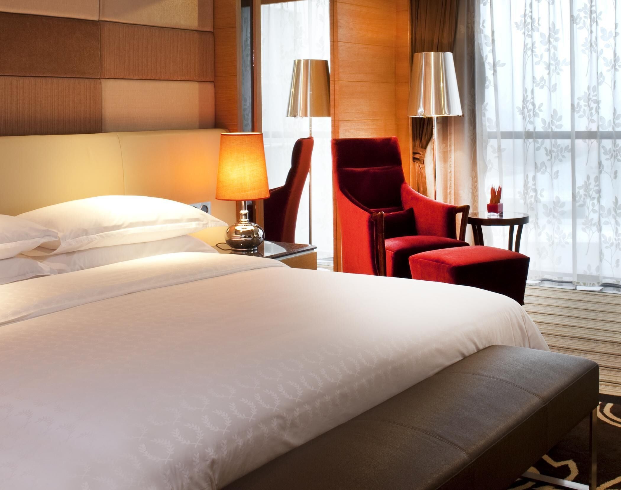 A hotel room with a large bed, two bedside lamps, a red armchair, and a small table with a vase.