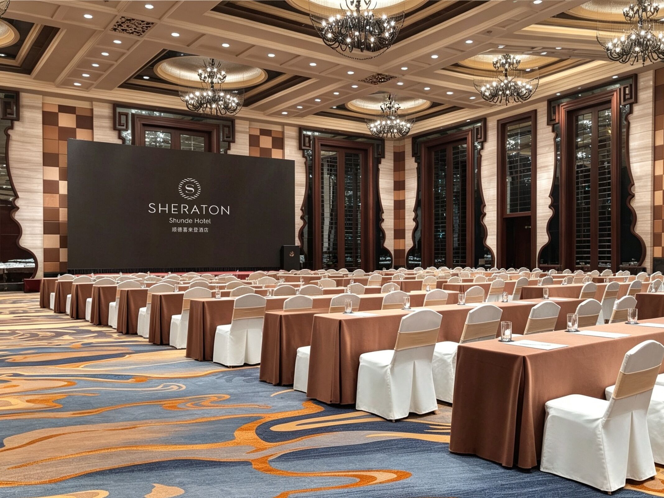 Salón de conferencias de un hotel Sheraton con filas de mesas, sillas blancas y una gran pantalla mostrando el nombre del hotel.