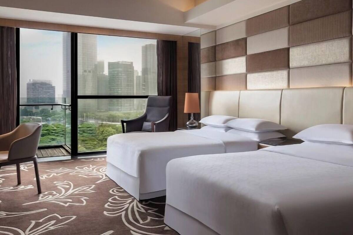 A hotel room with two beds, a chair, a lamp, and a large window offering a city view.