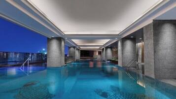 A large indoor swimming pool with clear water, surrounded by walls and columns, and a view of the city at dusk.