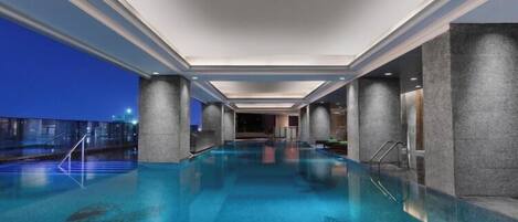 A large indoor swimming pool with clear water, surrounded by walls and columns, and a view of the city at dusk.