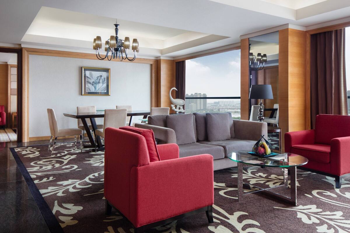 A modern hotel room with a living area featuring a sofa, armchairs, and a glass coffee table. A dining table with chairs is visible in the background.