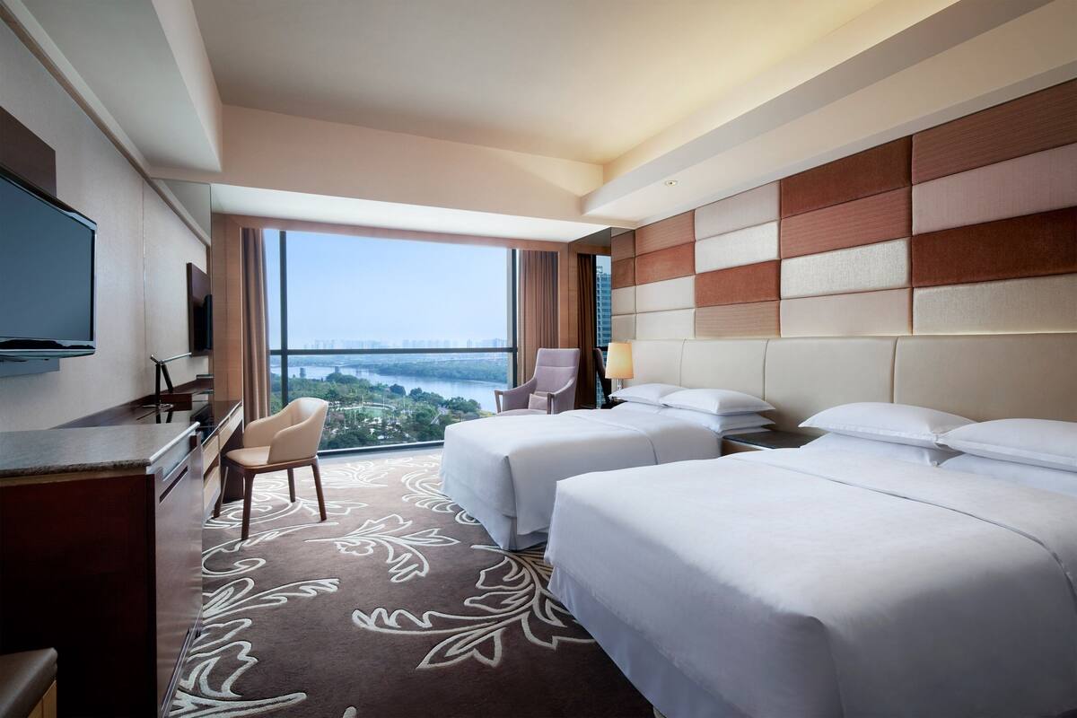 A hotel room with two beds, a desk, a chair, a TV, and a large window offering a city view.