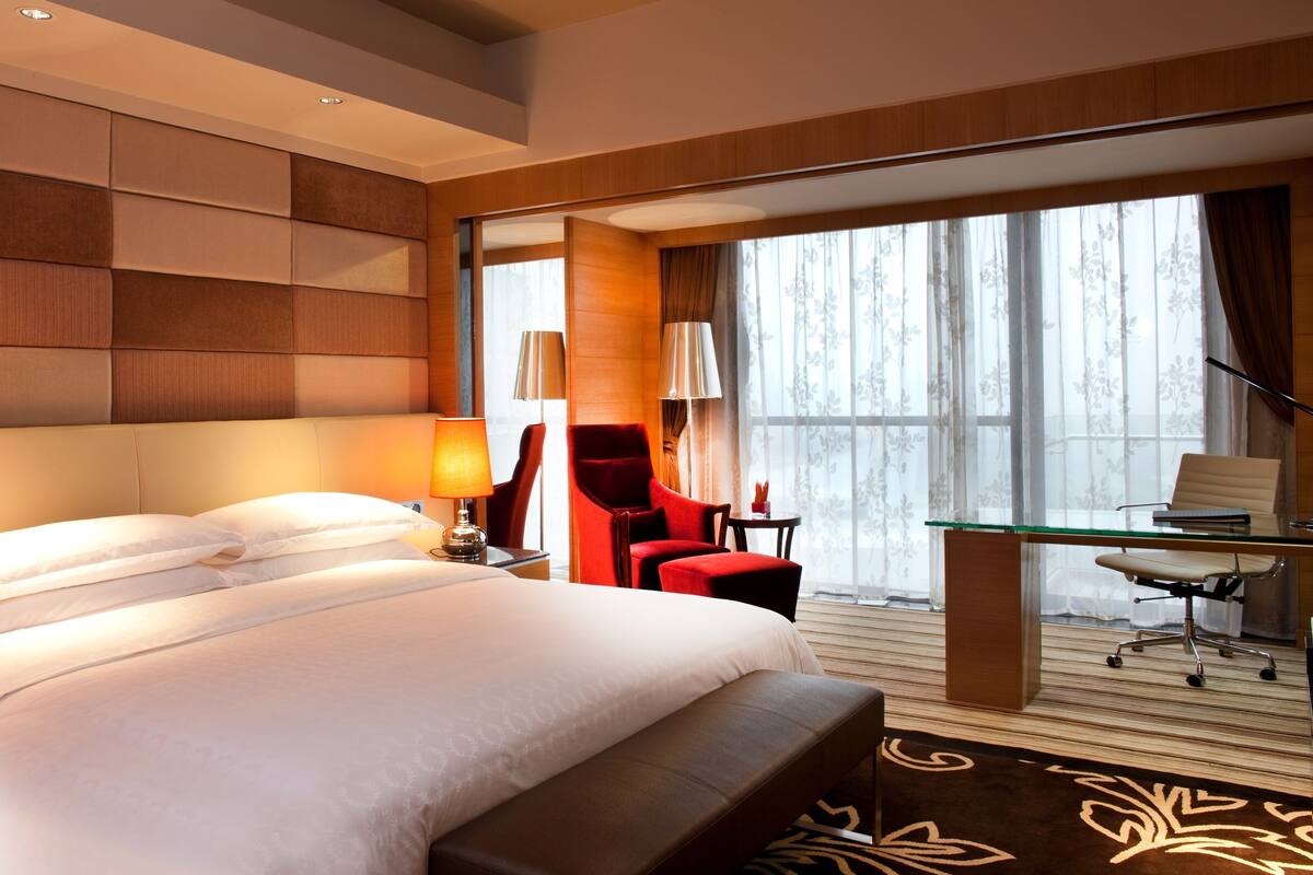 A hotel room with a large bed, a desk with a chair, a red armchair, and a view of the outdoors through a large window.