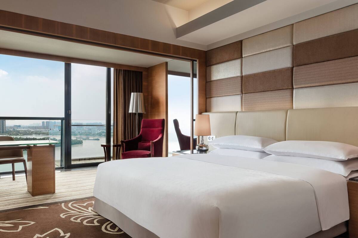 A hotel room with a large bed, a desk, a chair, and a view of a cityscape through large windows.