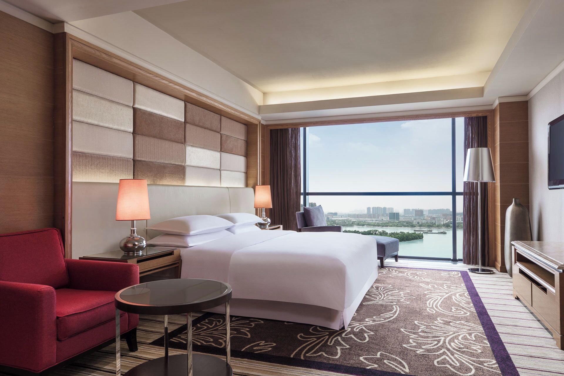 Quarto de hotel com uma cama grande, uma poltrona vermelha, uma mesa pequena e uma televisão.