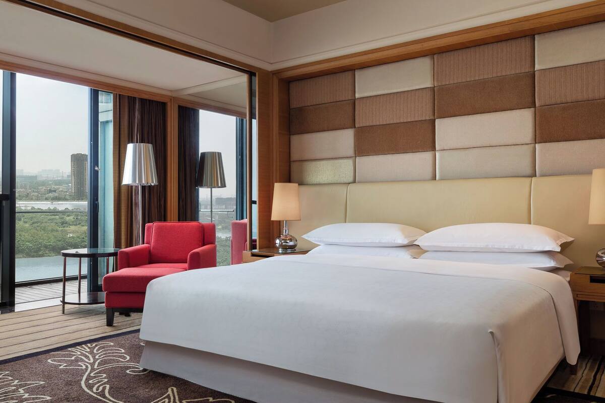 A hotel room with a large bed, a red armchair, a small table, and a view of a cityscape through large windows.