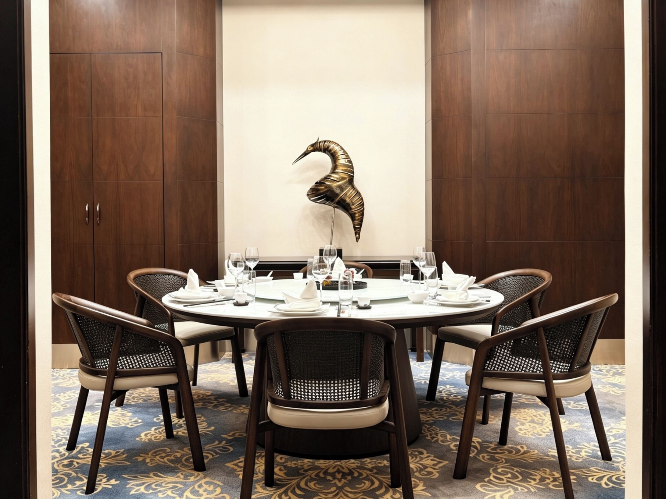 A dining area with a round table set for six, surrounded by wicker chairs, with a decorative metal sculpture on the wall.