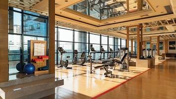 A spacious gym with various exercise equipment, large windows, and a wooden floor.