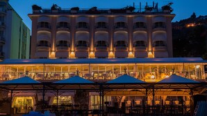 Ein Hotel mit mehreren Balkonen, Sitzgelegenheiten im Freien unter Markisen und einer großzügigen Fassade mit erleuchteten Fenstern.