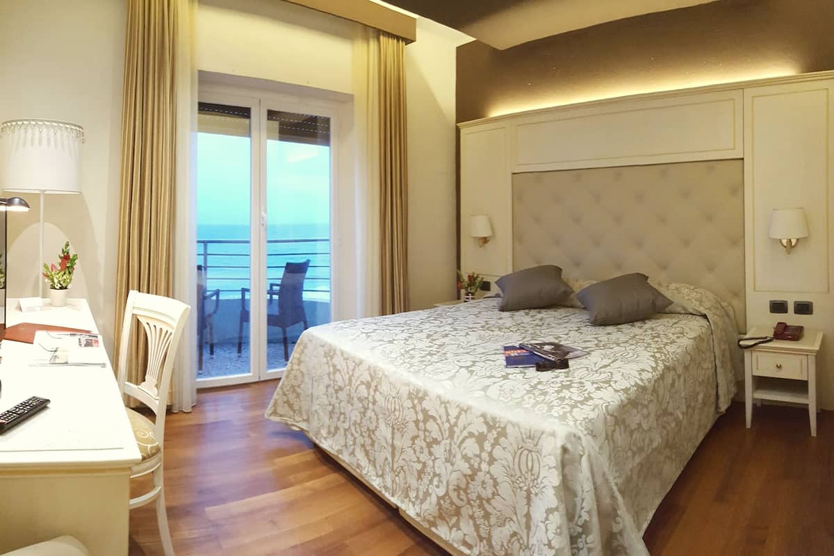 A hotel room with a bed, a desk, a chair, a nightstand, and a view of the sea through a sliding door.