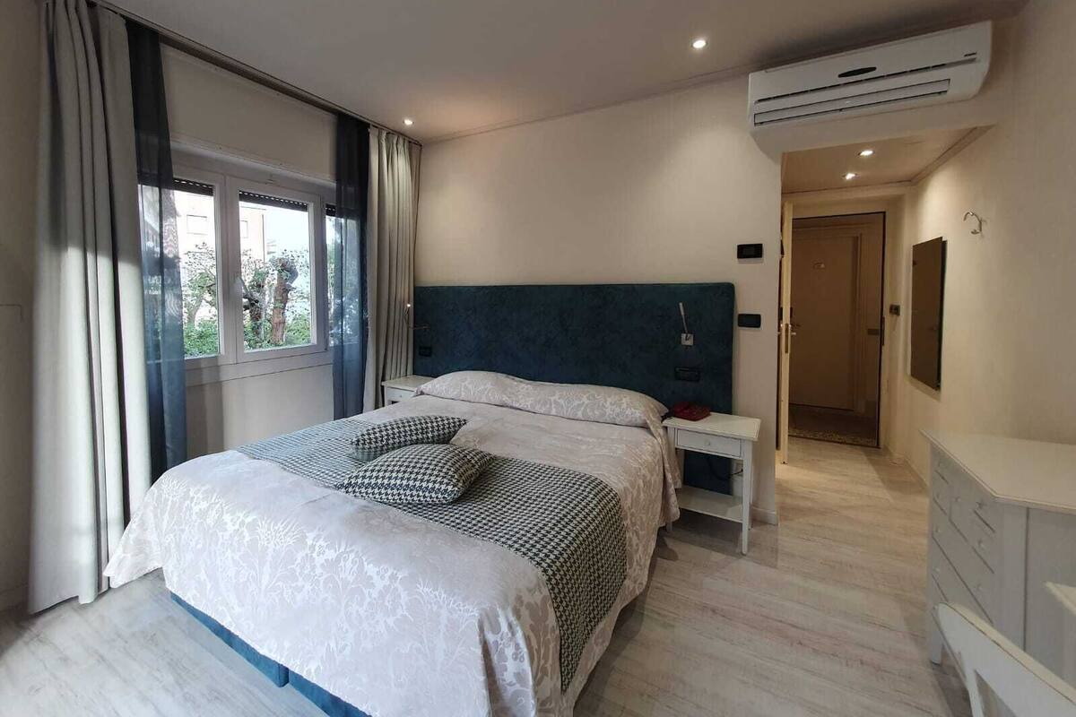 A modern bedroom with a large bed, a nightstand, an air conditioning unit, and a view of the outdoors through a window.