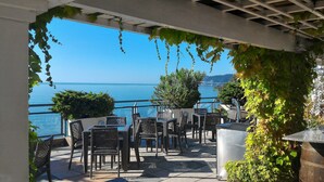 Eine Terrasse im Freien mit Korbmöbeln, Blick auf das Meer und viel Grün.