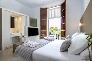 Habitación de hotel con cama, televisión, escritorio con espejo y ventana con contraventanas de madera.
