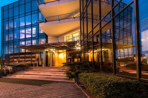 Een modern hotel met een glazen gevel, een centrale ingang met treden en een goed verlicht interieur.