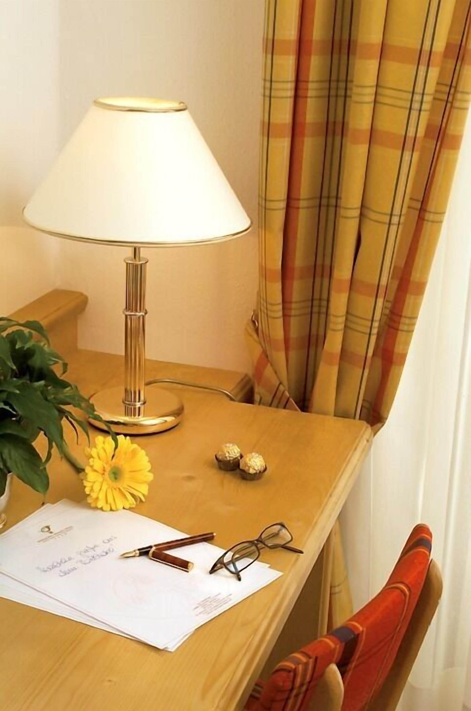 Una scrivania in legno con una lampada, un fiore giallo, una penna, occhiali e un foglio con scritto qualcosa.