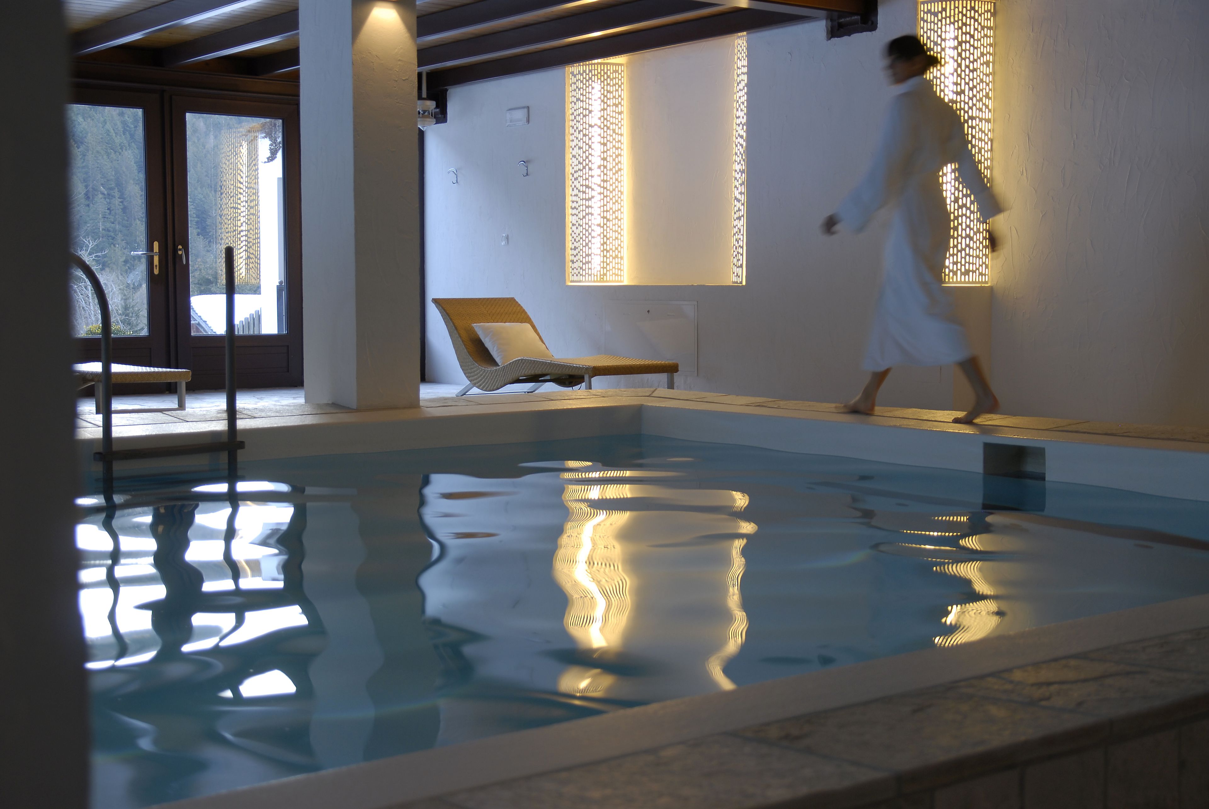 An indoor swimming pool with a wooden lounge chair and a person in a robe walking by.