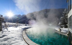 Ein Pool, umgeben von Schnee, mit aufsteigendem Dampf, ein Berg im Hintergrund und ein klarer Himmel.