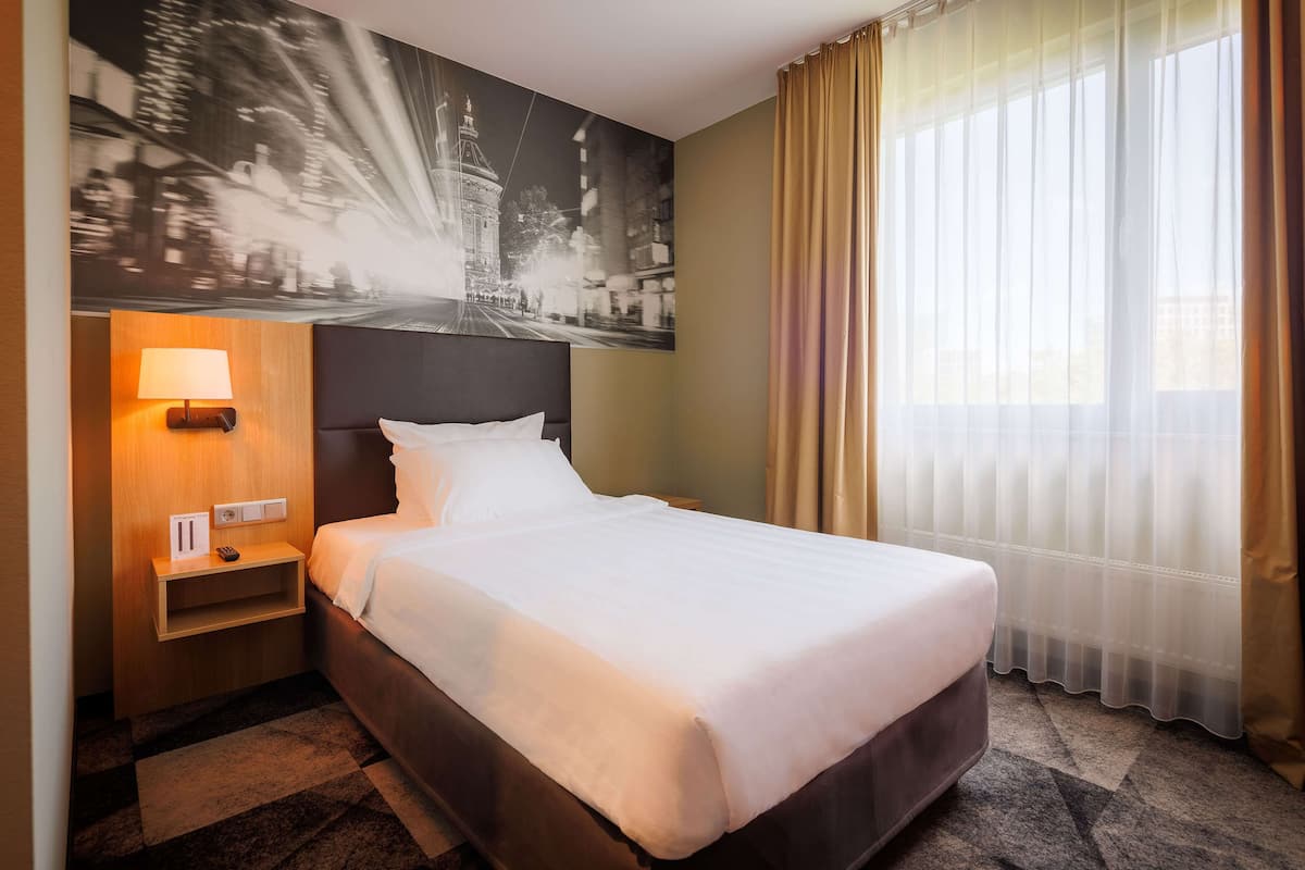 Una habitación de hotel con una cama grande, una mesita de noche con lámpara, un estante en la pared y una ventana con cortinas translúcidas.