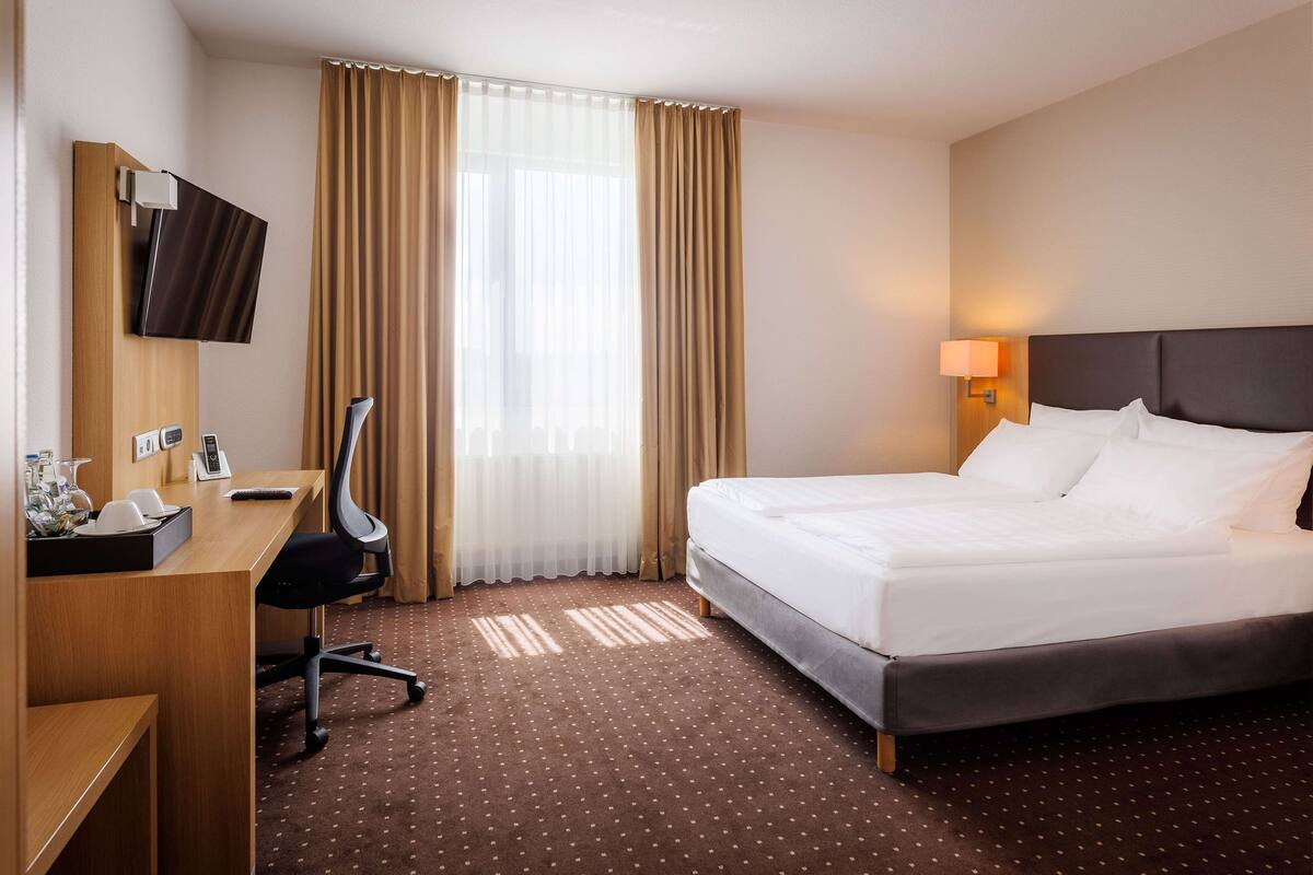 Una habitación de hotel con cama, escritorio, silla, televisión y ventana con cortinas.