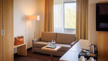 A hotel room with a sofa, a small table with a tray of glasses and a bottle, a coffee maker, and a TV on the wall.