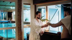 Un homme, vêtu d’un peignoir blanc, se tient près d’une fenêtre d’où l’on aperçoit une piscine.