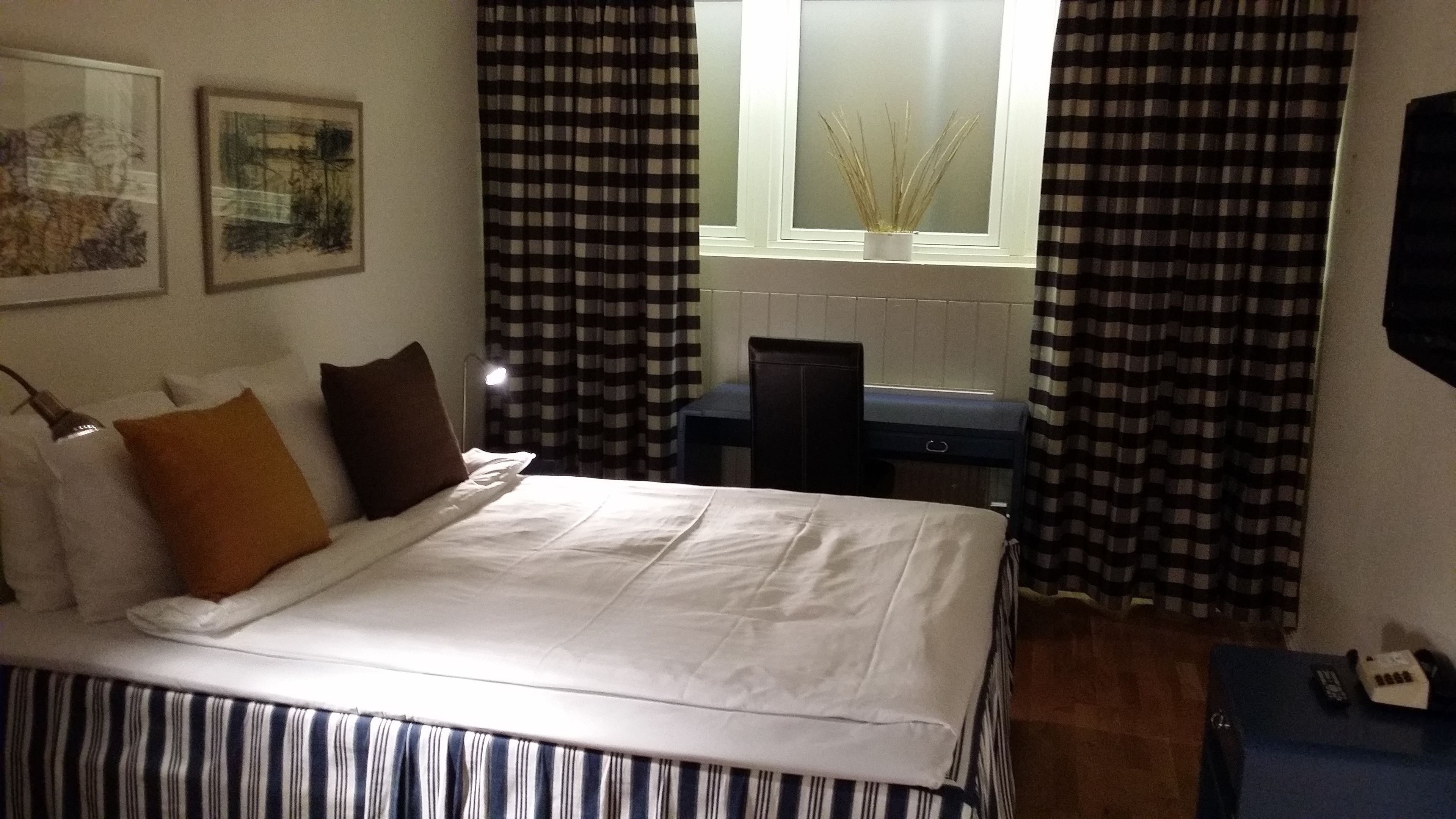 Un lit bien fait, avec du linge de lit blanc, un couvre-lit à rayures et deux oreillers. On y trouve un petit bureau avec une chaise et une lampe, une fenêtre donnant sur l’extérieur et un téléviseur fixé au mur.